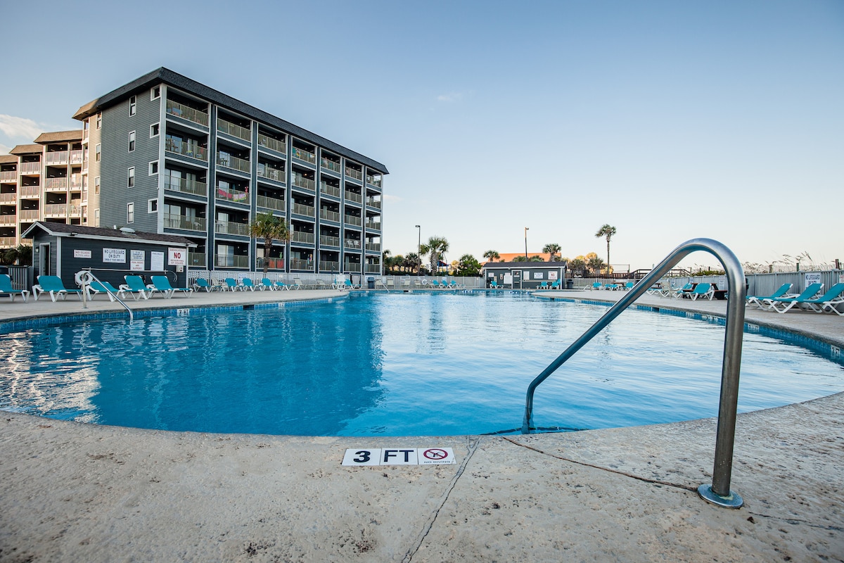 The outdoor pool area is surrounded by lounge chairs and features crystal-clear water, with a gentle slope leading into the shallow section marked at 3 feet. A modern multi-story building can be seen in the background against a clear blue sky.