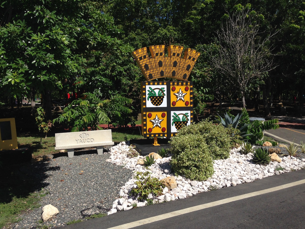 A colorful artistic structure representing Yauco is surrounded by lush greenery and well-maintained gravel pathways. A stone bench is positioned nearby, offering a resting spot for visitors. Various plants, including succulents, are arranged harmoniously in the landscape.