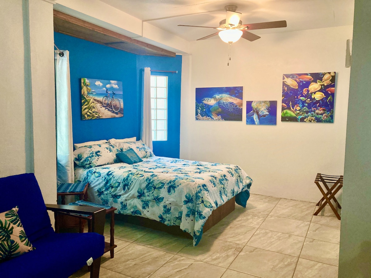 A serene sleeping area features light blue walls adorned with ocean-themed artwork. A spacious bed is dressed in floral-patterned linens, complemented by a cozy blue chair and a small folding table. Natural light enters through a window, accentuated by light curtains.