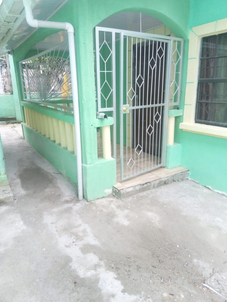 An entrance features a gated screen door, providing security and access to the interior. Bright green walls complement the architecture, while the adjacent area includes a covered porch with a railing. Natural light is present through the nearby windows.