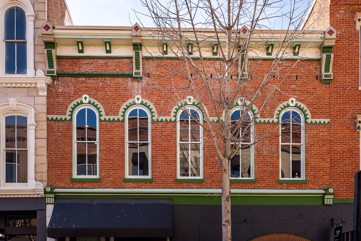 The exterior of a historic brick building is displayed, featuring five tall, arched windows with green trim. The facade exhibits intricate detailing above the windows and contrasting black elements at the base. A bare tree is seen in front, adding to the urban landscape.