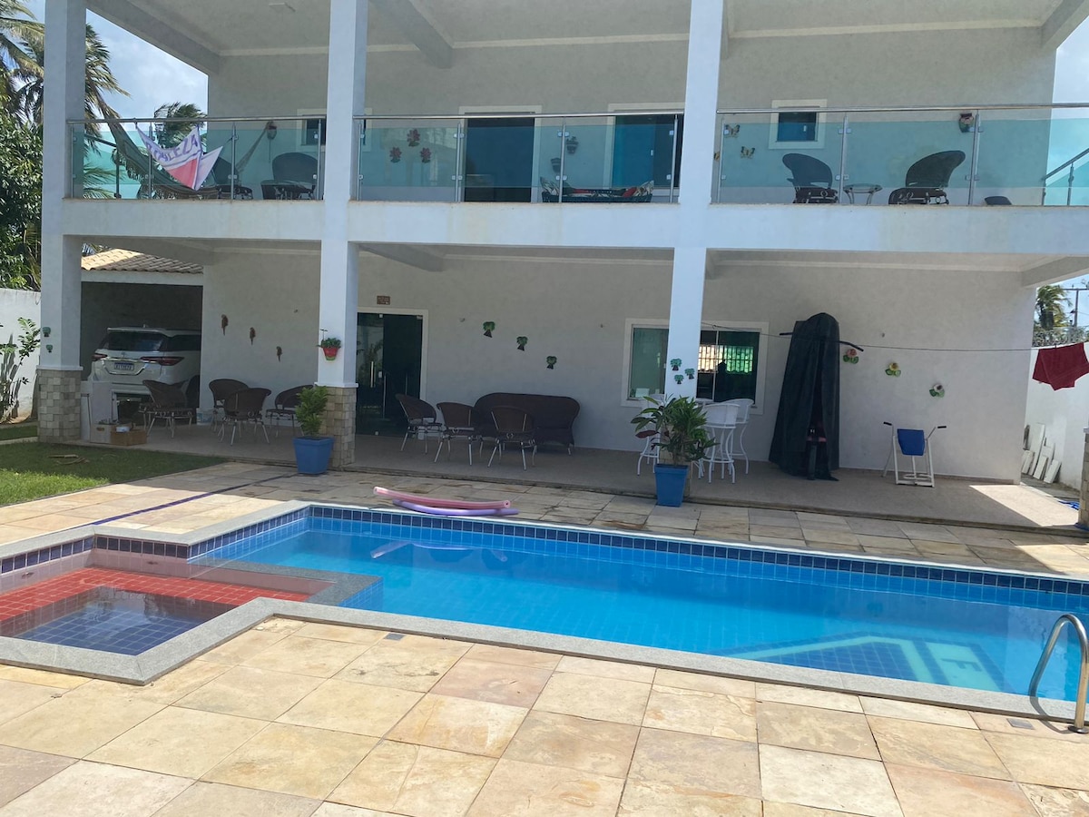 A spacious outdoor area features a rectangular swimming pool surrounded by a tiled patio. Lounge chairs are arranged near the pool, with a small section visible for seating. Two levels of the building above show a balcony with multiple chairs and potted plants, creating a relaxed environment.