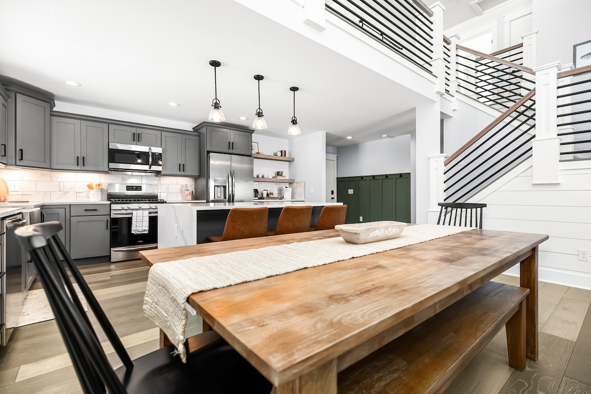 A modern kitchen and dining area is showcased, featuring sleek gray cabinetry and stainless steel appliances. A long wooden dining table is centered, accented by black chairs. Pendant lights hang above, illuminating the space, while open shelving displays decorative items.