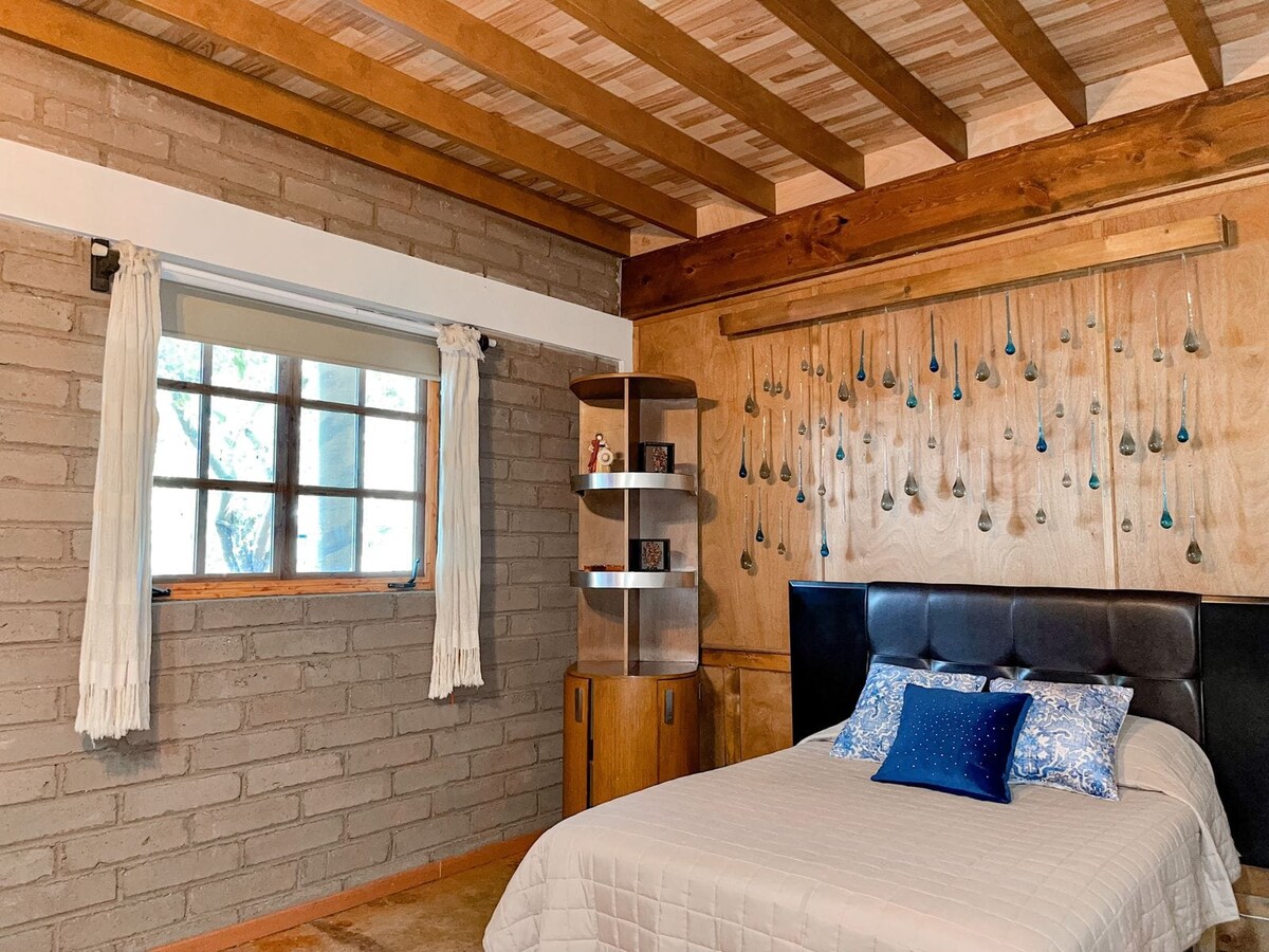 A cozy sleeping area features a spacious bed with a quilted cover and decorative pillows. A wooden shelf with decorative items is positioned beside the bed. Natural light enters through a window framed by lightly colored curtains, enhancing the simplicity of the rustic brick and wooden wall design.