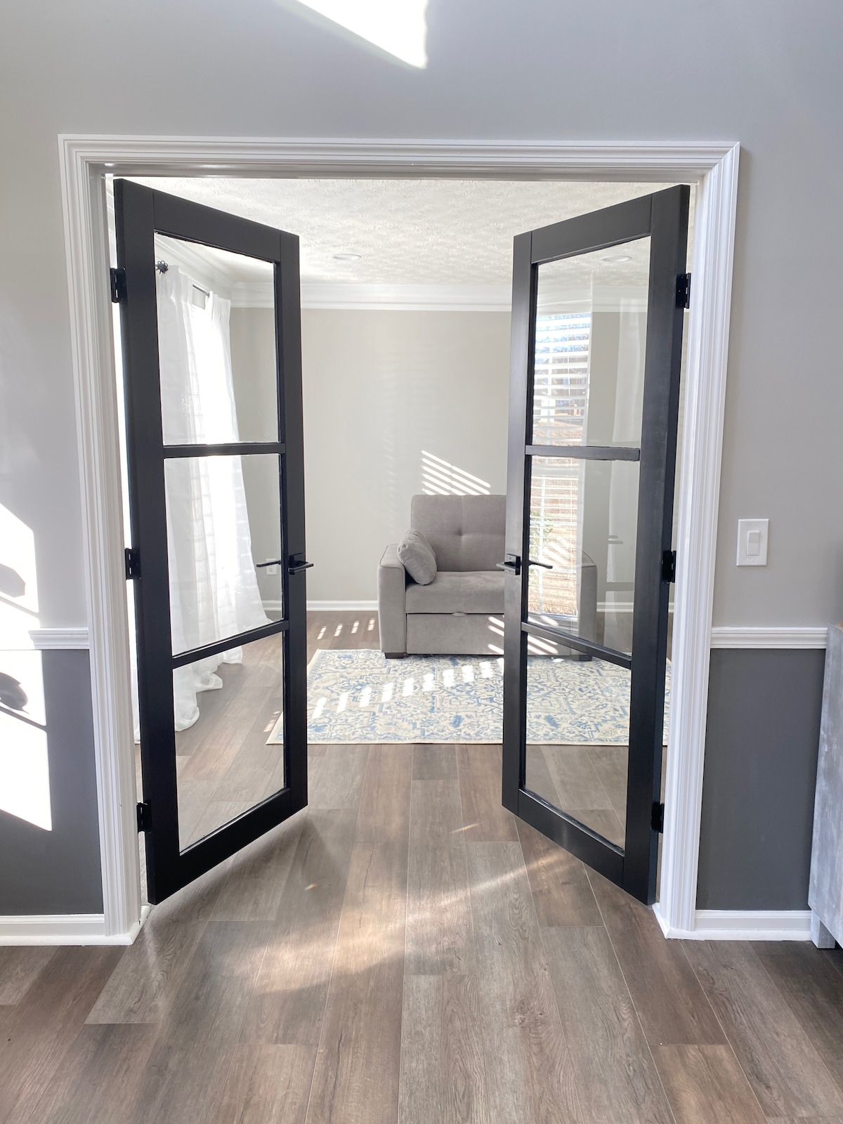 Double glass doors open into a well-lit room featuring a cozy gray chair. Sunlight streams through the partially drawn sheer curtains, casting soft patterns on the floor. A patterned area rug is visible beneath the chair, contributing to the room's inviting atmosphere.