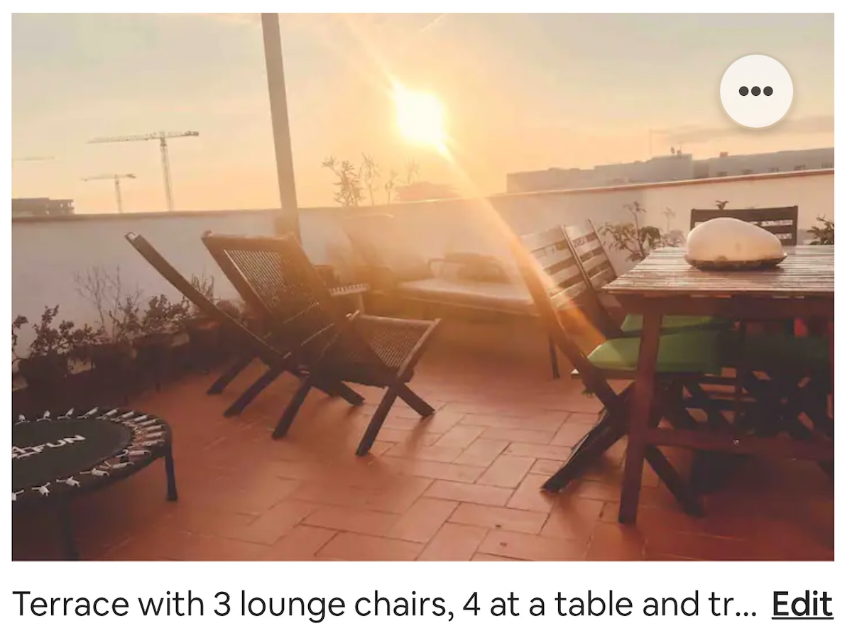 A spacious terrace is shown, featuring three lounge chairs and a table with seating for four. The setting sun casts a warm glow over the area, illuminating the terracotta tiles and surrounding plants.