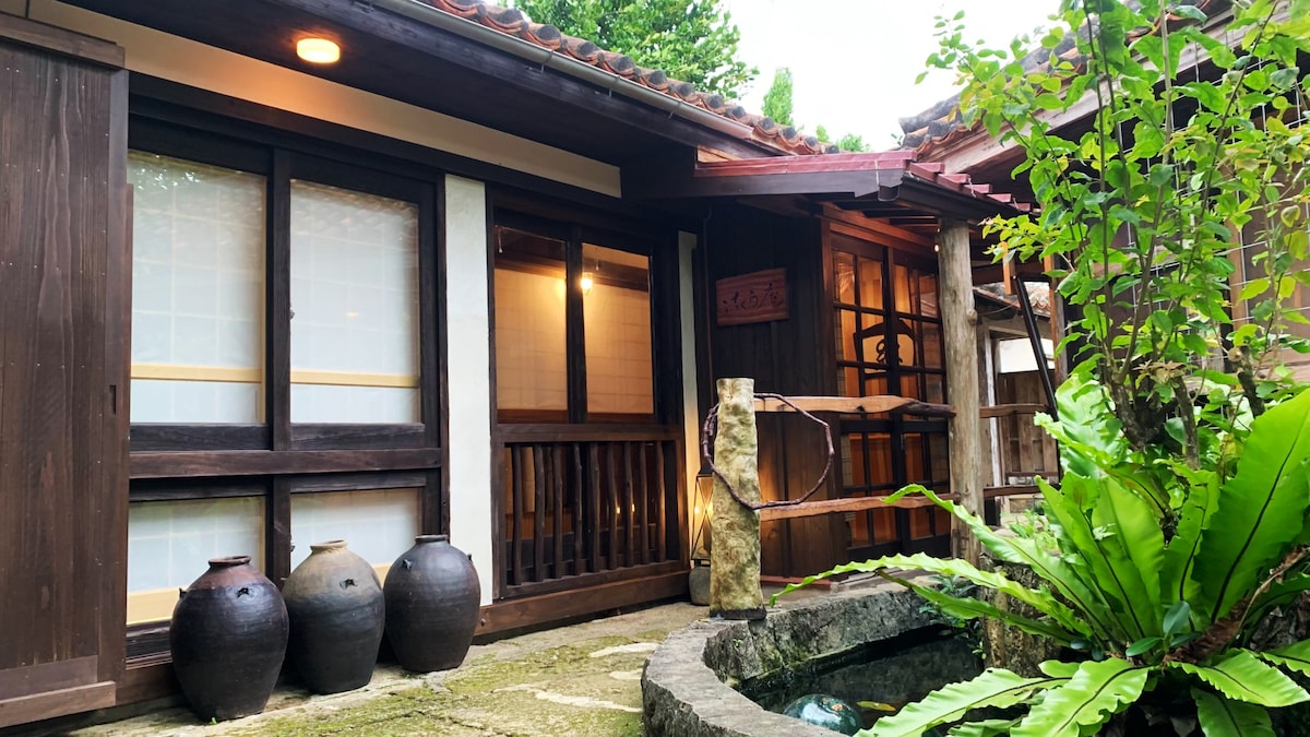 A traditional Japanese-style exterior is presented, featuring wooden beams and sliding doors. A small stone water feature is surrounded by lush greenery, including large ferns. Several clay pots are placed near the entrance, enhancing the serene and nature-inspired ambiance.