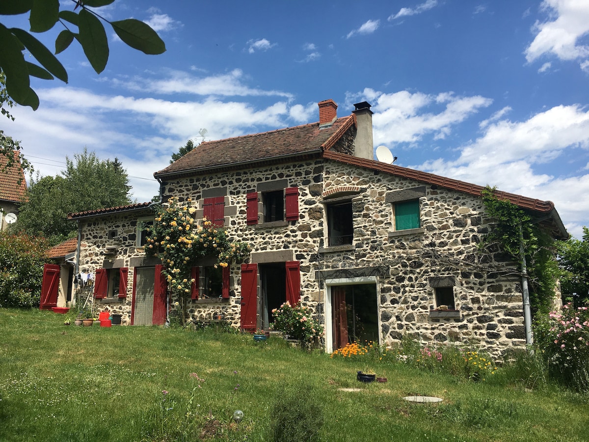 A charming stone house is set within a lush green landscape. Red window shutters contrast with the natural stone exterior. Flowering plants and manicured grass surround the home, while a clear blue sky with scattered clouds provides a serene backdrop.