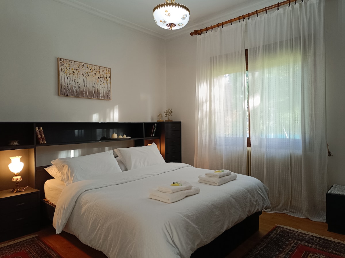 A spacious bedroom features a large bed with white linens and neatly arranged towels. A decorative artwork hangs above the bed, while soft light filters through sheer curtains, illuminating the warm wooden floors.