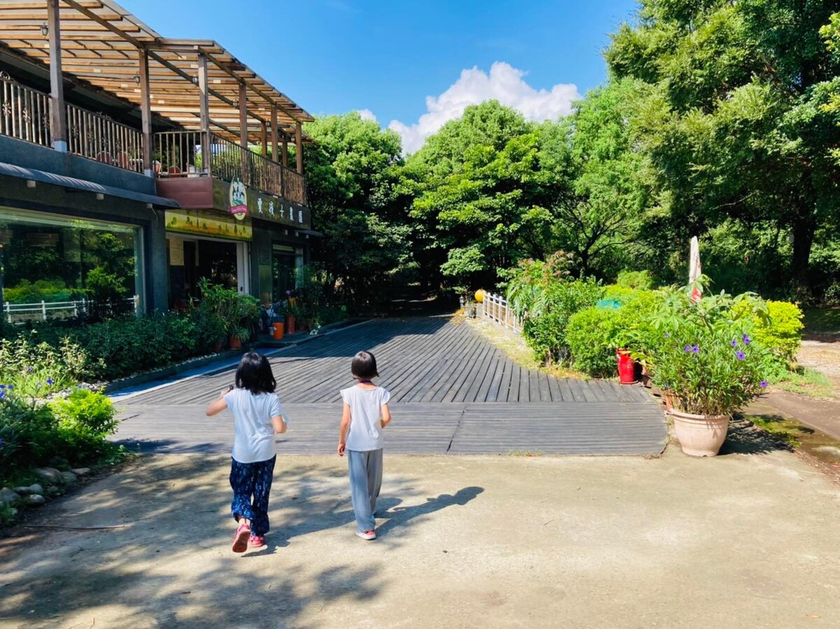 A wooden walkway is seen leading towards a green area, framed by trees and colorful plants. Two children walk side by side on the path, enjoying the serene outdoor environment. The sky is clear, with a few clouds visible in the background.
