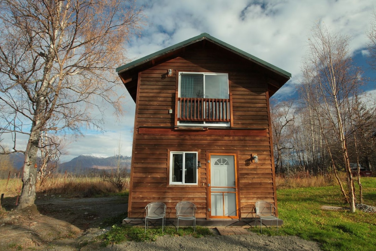 The wooden cabin features a two-story design with a welcoming entrance. A small balcony is visible above the front door, and four chairs are positioned on the ground level in a grassy area. Surrounding trees and scenic mountain views are present in the background.