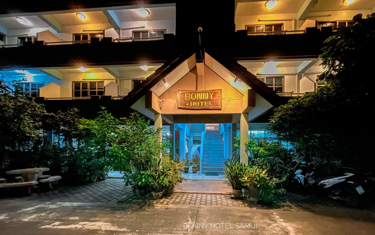 The entrance to Bonny Hotel is framed by lush greenery, showcasing a warm, welcoming facade. Soft lighting highlights the hotel name and the structure's contemporary design. The stairway leads to upper levels, while the surrounding area features a mix of stone pathways and greenery.