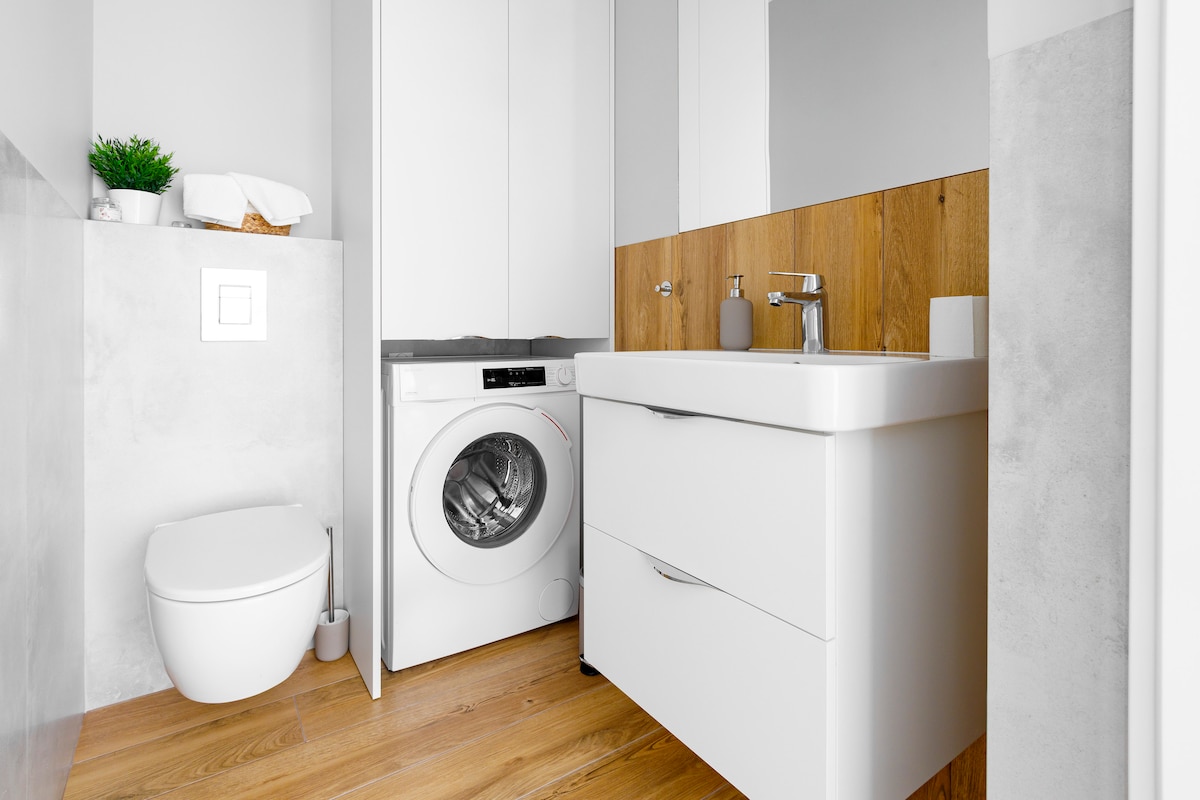 A modern bathroom features minimalist design with wood accent walls. A washing machine is situated next to a sleek white sink with a matching cabinet. A toilet is located adjacent, and natural light brightens the space, complemented by white towels and decorative greenery.