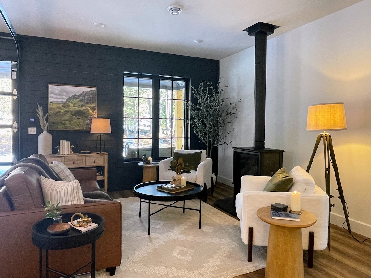 A cozy living room features a warm gas fireplace and two inviting armchairs opposite a comfortable sofa. A circular coffee table is centered on a textured rug, complemented by a floor lamp and a side table. Large windows allow natural light to illuminate the space.