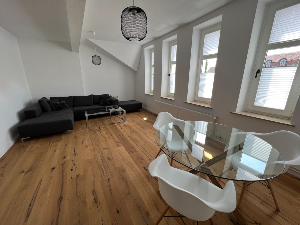 A living space is presented with a sectional sofa in a neutral tone and a glass coffee table. Natural light filters through multiple windows, highlighting the light wood flooring and white walls. A round dining table with modern chairs is visible, adding to the open layout.