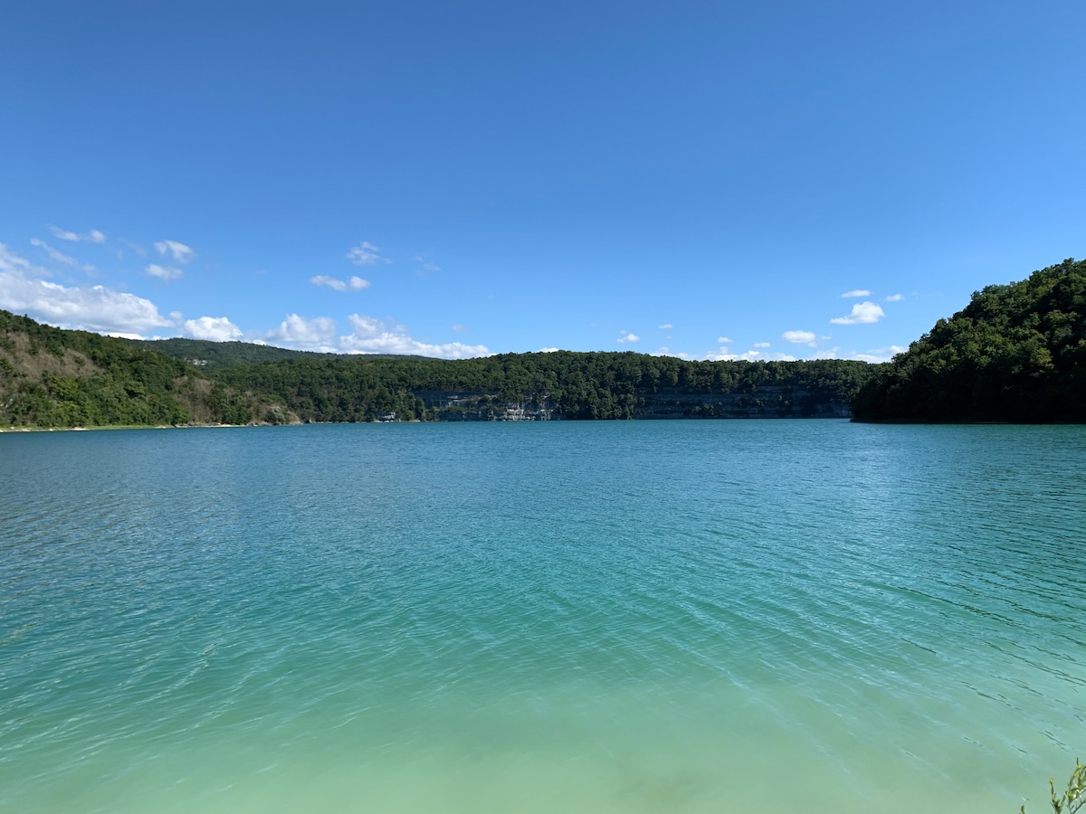 A serene lake is displayed, featuring clear turquoise waters that gradually transition to a lighter hue near the shore. Lush greenery lines the banks, while distant cliffs and a blue sky with scattered clouds create a peaceful backdrop.