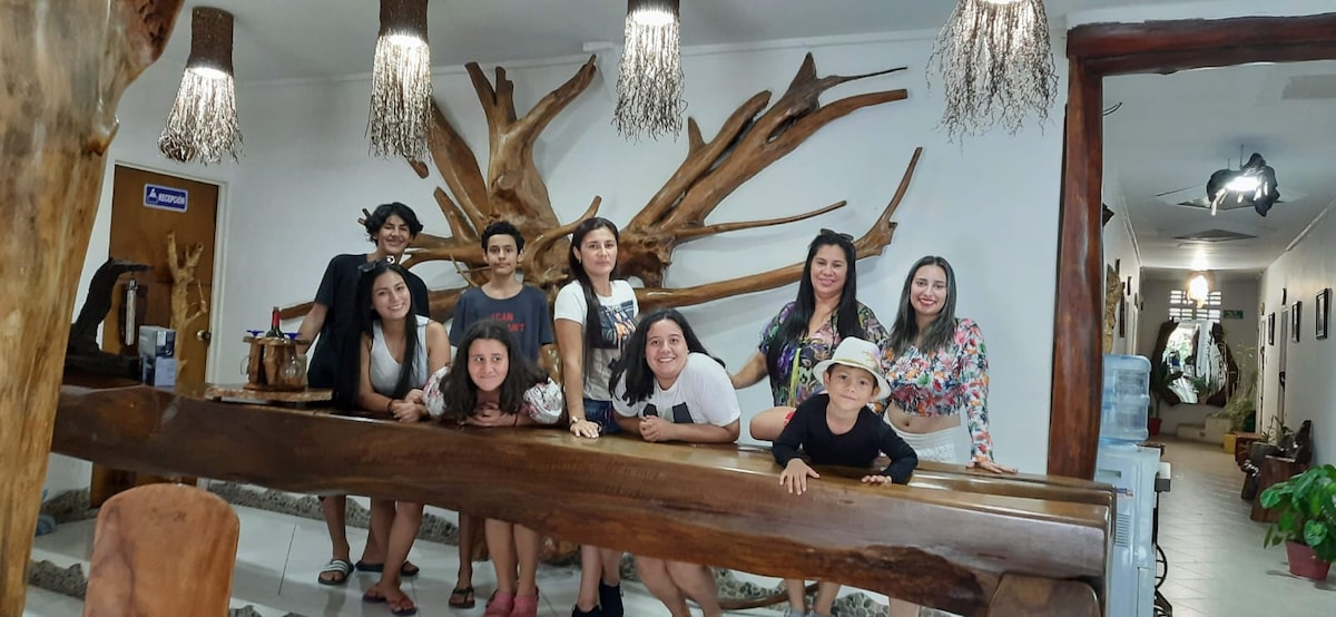 A long wooden reception desk is highlighted, surrounded by artistic wooden decorations on the wall. Several individuals gather around the desk, engaging with one another in a warm atmosphere. The interior is well-lit, with decorative lighting fixtures hanging from the ceiling.