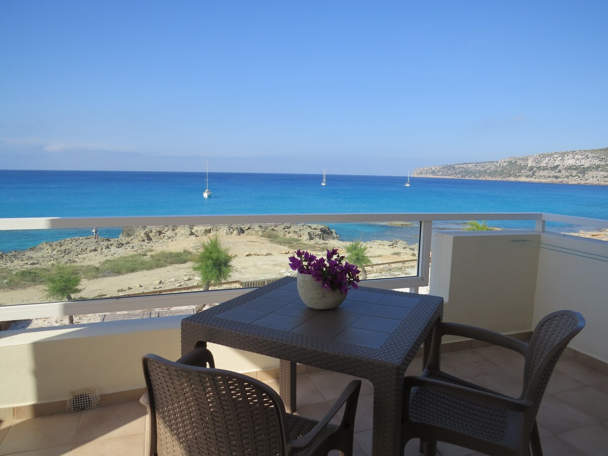 A terrace features a table with four chairs, accompanied by a planter filled with purple flowers. Scenic views of the clear blue ocean and distant sailboats can be observed, framed by the rocky shoreline under a bright blue sky.