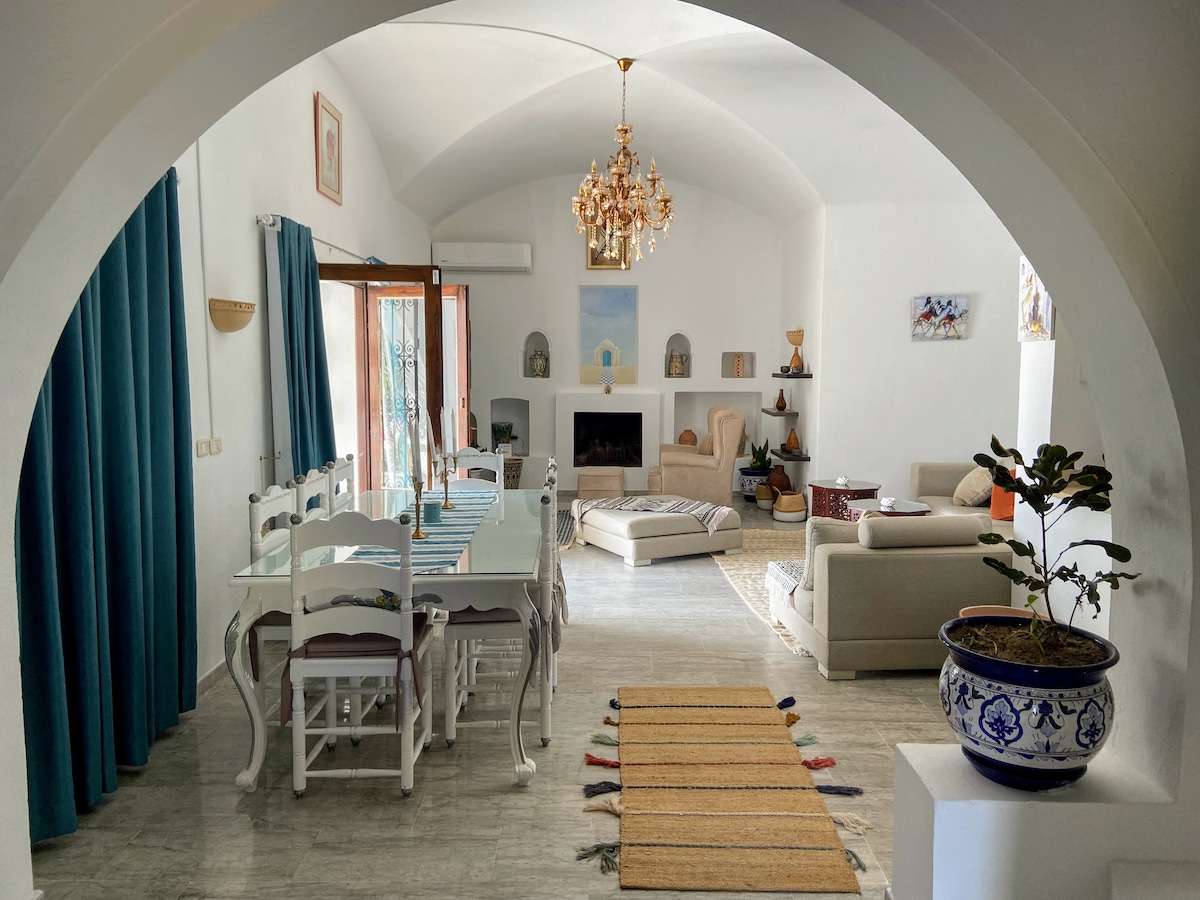 A spacious living area is presented, featuring an elegant chandelier and large windows that let in natural light. A dining table with multiple chairs is positioned alongside a comfortable seating arrangement. Decorative elements, including a modern fireplace and potted plants, enhance the inviting ambiance.
