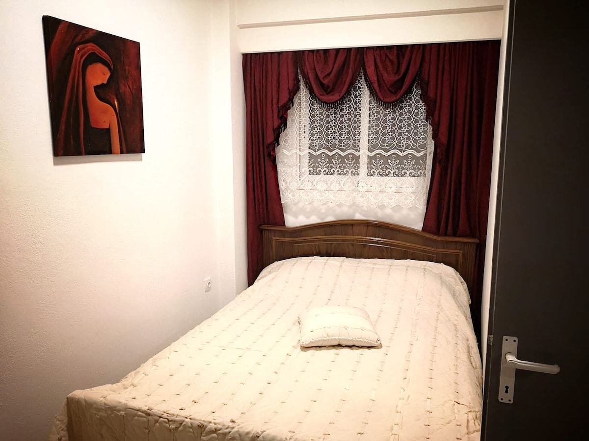 A cozy sleeping area features a double bed with a light-colored coverlet. Rich red drapes frame a window adorned with lace curtains. Abstract artwork is displayed on the wall, providing a touch of character to the space.