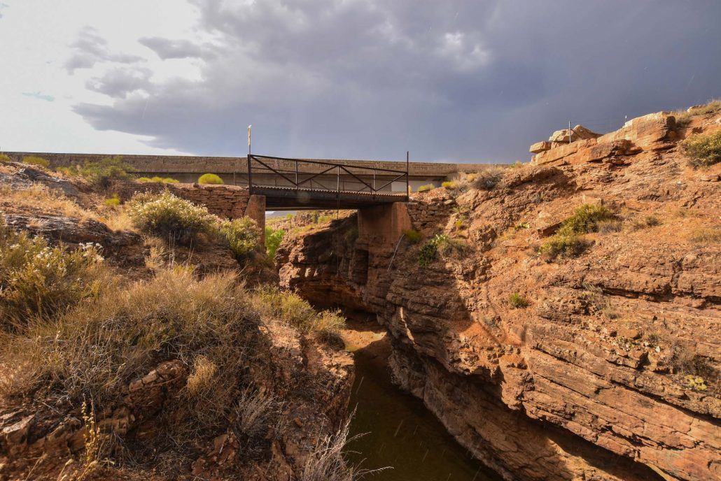 A sturdy metal bridge spans a rocky canyon, connecting two rugged landforms. Surrounding the bridge, dry grass and shrubs contrast against the exposed stone walls. The sky is overcast, hinting at an impending storm, while water is visible below the bridge, nestled within the natural landscape.