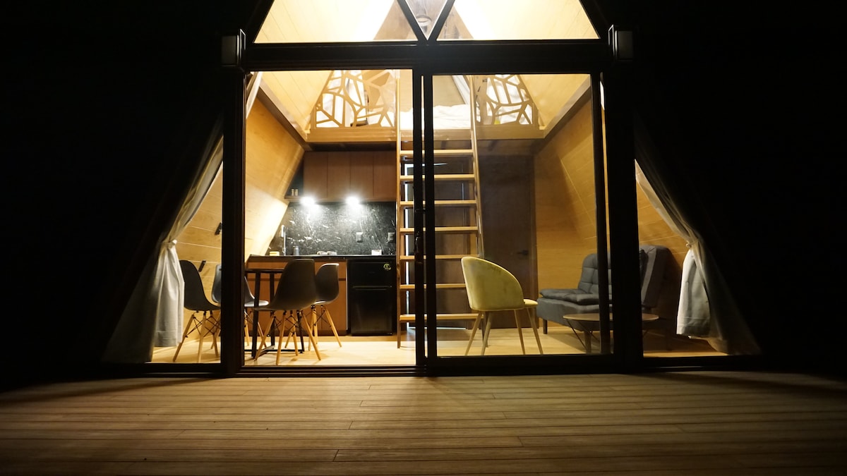 The A-frame cabin is showcased at night, displaying large glass doors that reveal a well-lit interior. Inside, a modern dining area is seen, complemented by stylish seating and a staircase leading to the upper sleeping loft, all illuminated by soft overhead lighting.