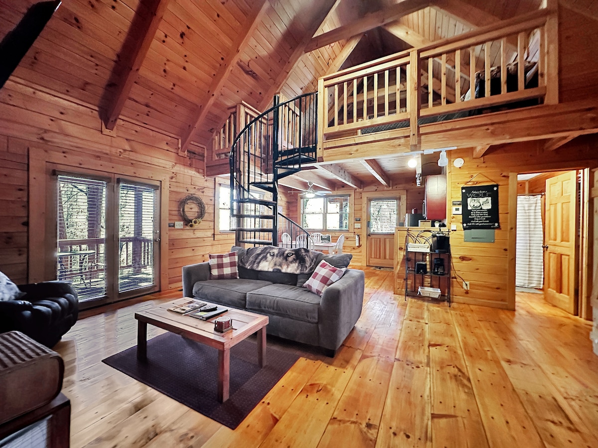 An open-concept living area showcases natural wood paneling and flooring, complemented by a cozy sofa and a coffee table. A spiral staircase leads to a lofted sleeping area, while large windows provide views of the surrounding trees. The space is illuminated by soft, ambient lighting.