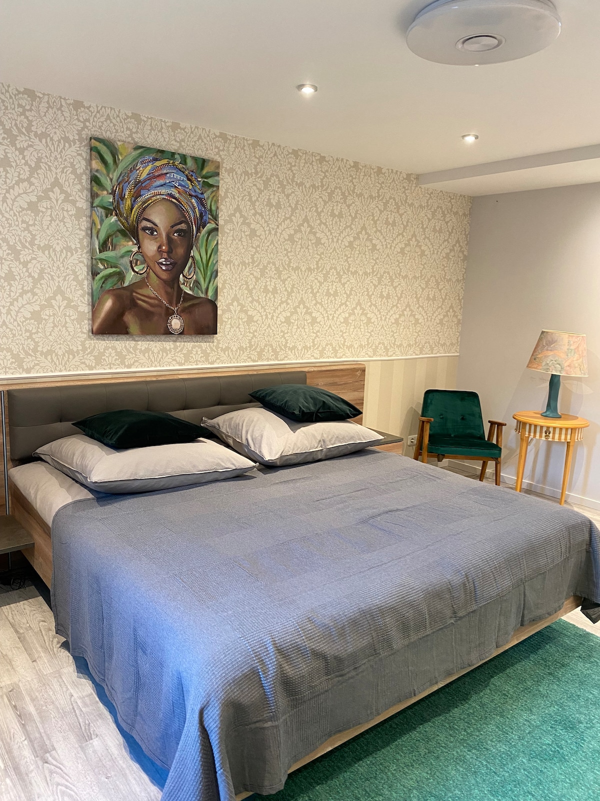 A spacious sleeping area features a comfortable bed with two pillows and a textured blanket. A vibrant piece of wall art is displayed above the bed, while a green armchair and a small wooden side table are situated nearby. Soft lighting is provided by recessed ceiling lights.