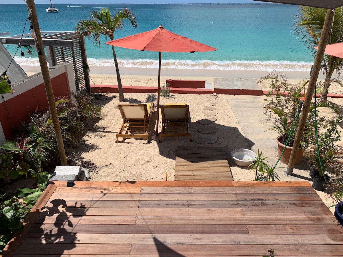 A sandy garden area features two beach chairs beneath red umbrellas, offering direct views of the calm sea and sandy beach. A wooden deck provides access to the space, while tropical plants enhance the inviting environment.