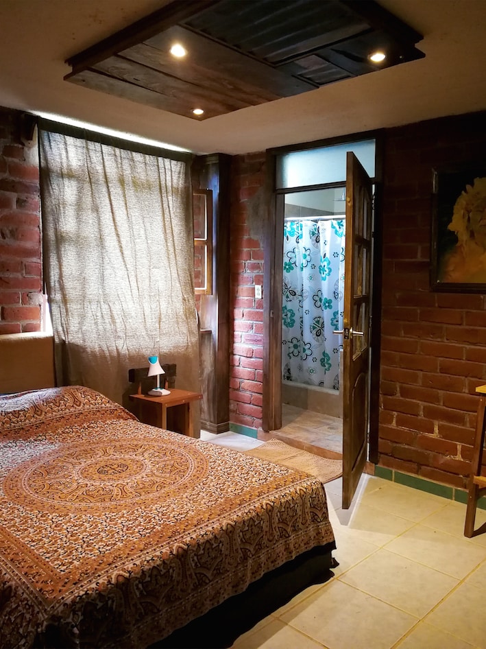 A bedroom features a spacious bed draped in a patterned blanket, complemented by a rustic wooden nightstand. Natural light filters through a partially closed curtain. A doorway leads to a bathroom area, visible in the background, adorned with patterned tiles.