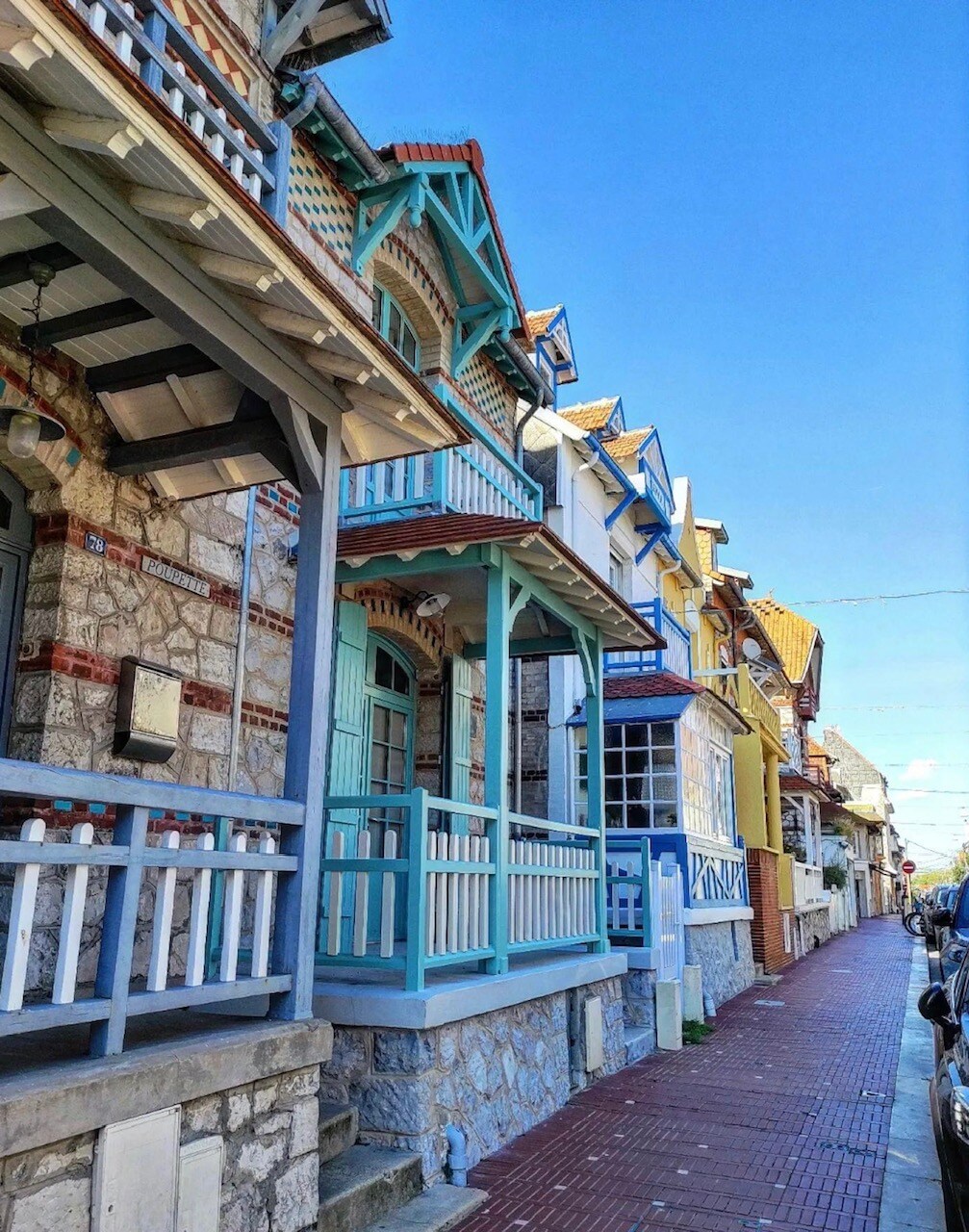 A row of charming houses is displayed, featuring unique architectural details and colorful facades. Balconies with decorative railings are visible, alongside stone and wood elements. A clear blue sky provides a bright backdrop, enhancing the inviting appearance of the neighborhood.