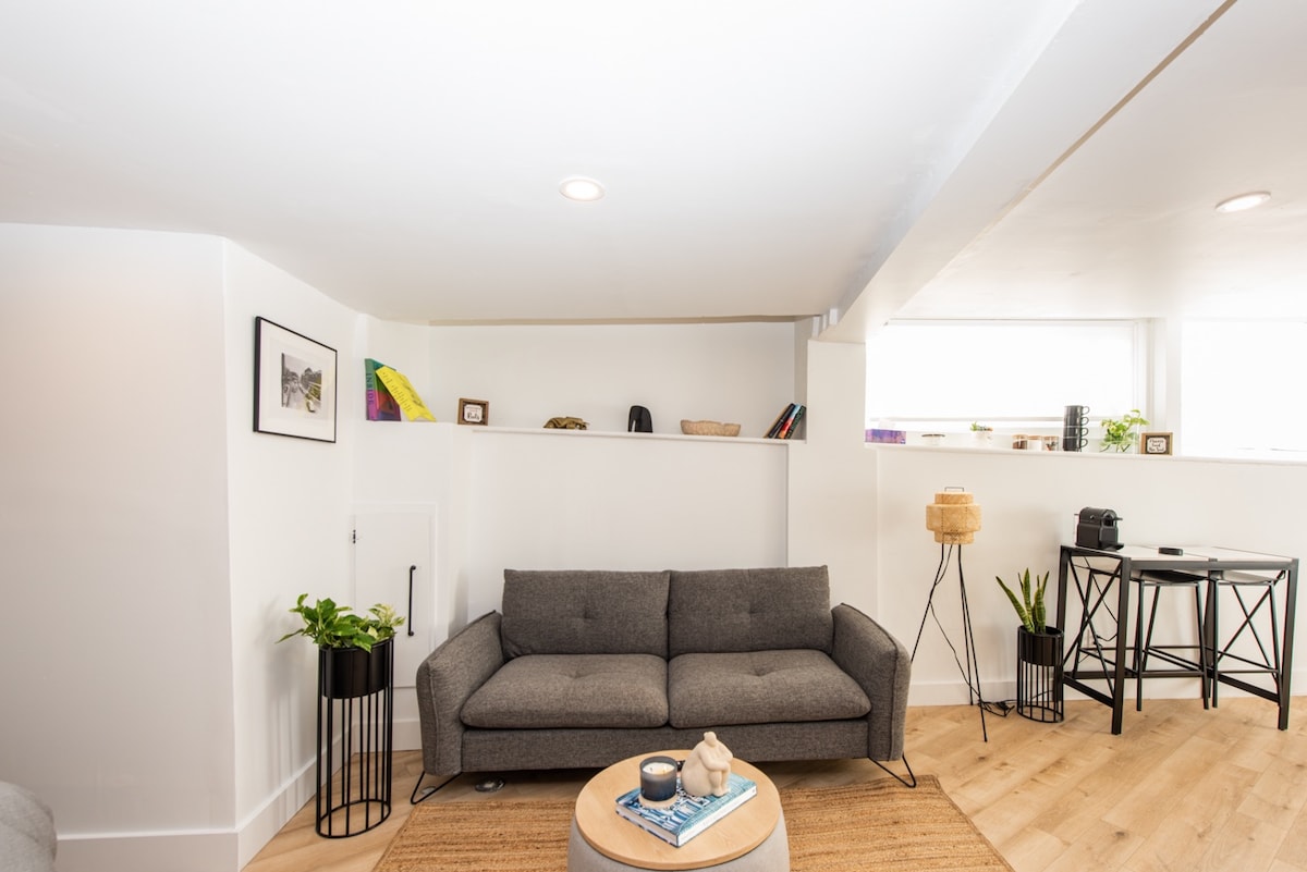 A modern living space features a gray sofa positioned beside a round coffee table. Shelves above offer decorative items and small plants, while a lamp and small table stand nearby. Light enters through large windows, illuminating the warm wood flooring.