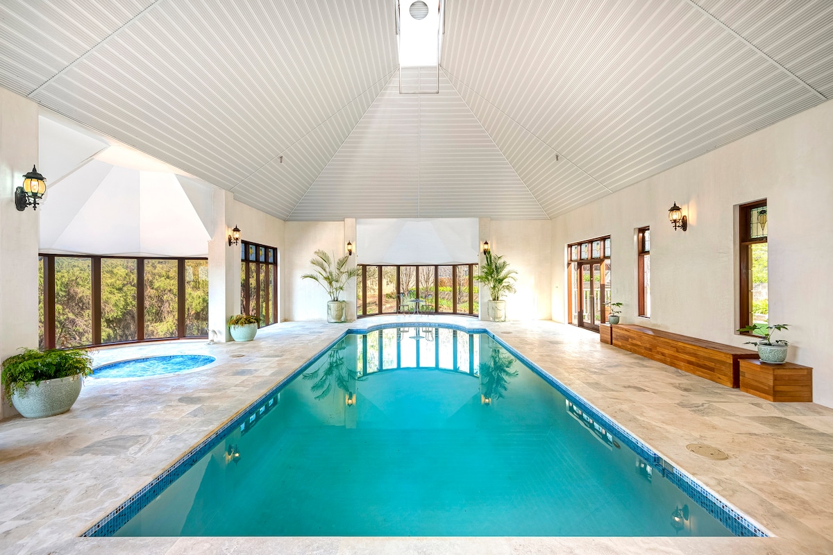 A spacious indoor pool area features a large, inviting pool surrounded by natural stone tiles. Tall glass windows offer serene views of the outdoors, while potted plants add a touch of greenery. Soft, neutral tones enhance the open and airy atmosphere.