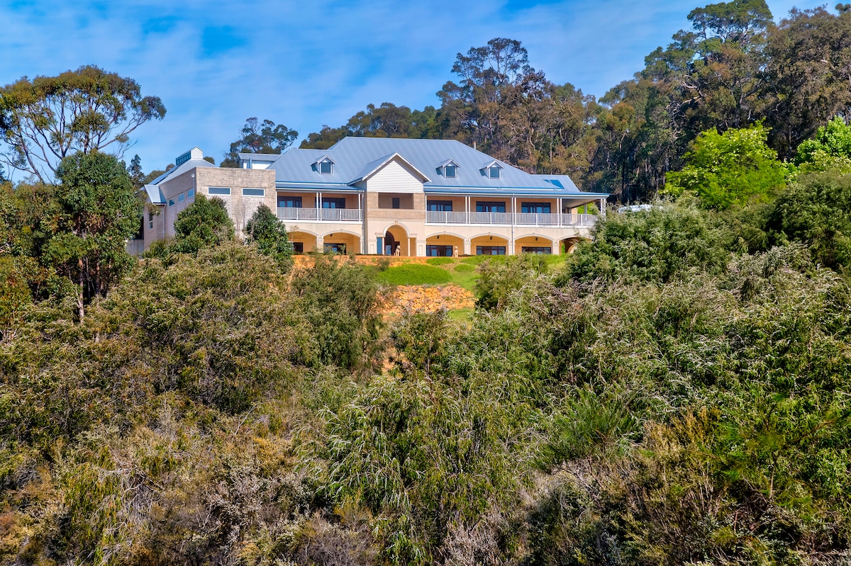 The multi-story building is set against a backdrop of lush greenery, featuring a modern design with a blue roof and multiple arched windows. The structure is surrounded by native Australian plants, providing a serene and natural environment.