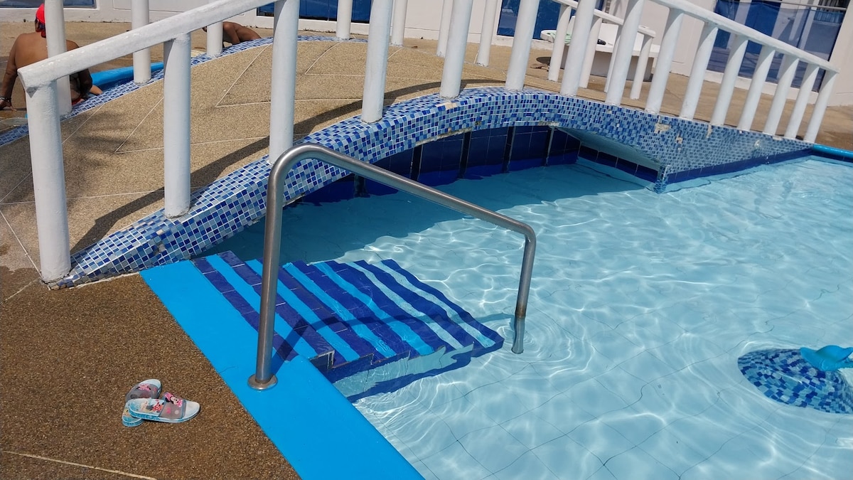 The image features a swimming pool with a gentle slope leading to the water. A metal handrail provides support next to the steps, which are adorned with striped tiles. The pool area includes a textured surface and a single flip-flop resting on the edge.