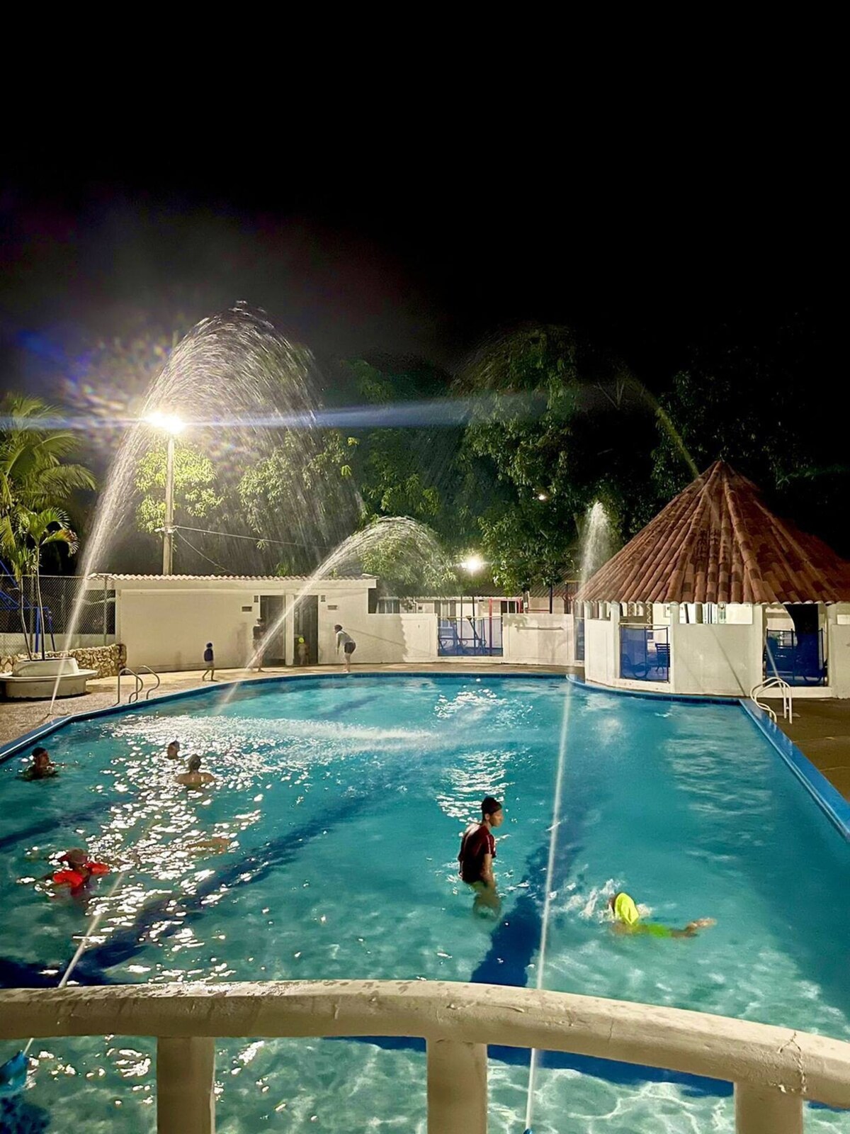 A swimming pool is illuminated at night, surrounded by water fountains that create gentle sprays. Several individuals enjoy the water, with palm trees visible in the background. A thatched-roof structure is present, adding to the relaxing ambiance of the area.