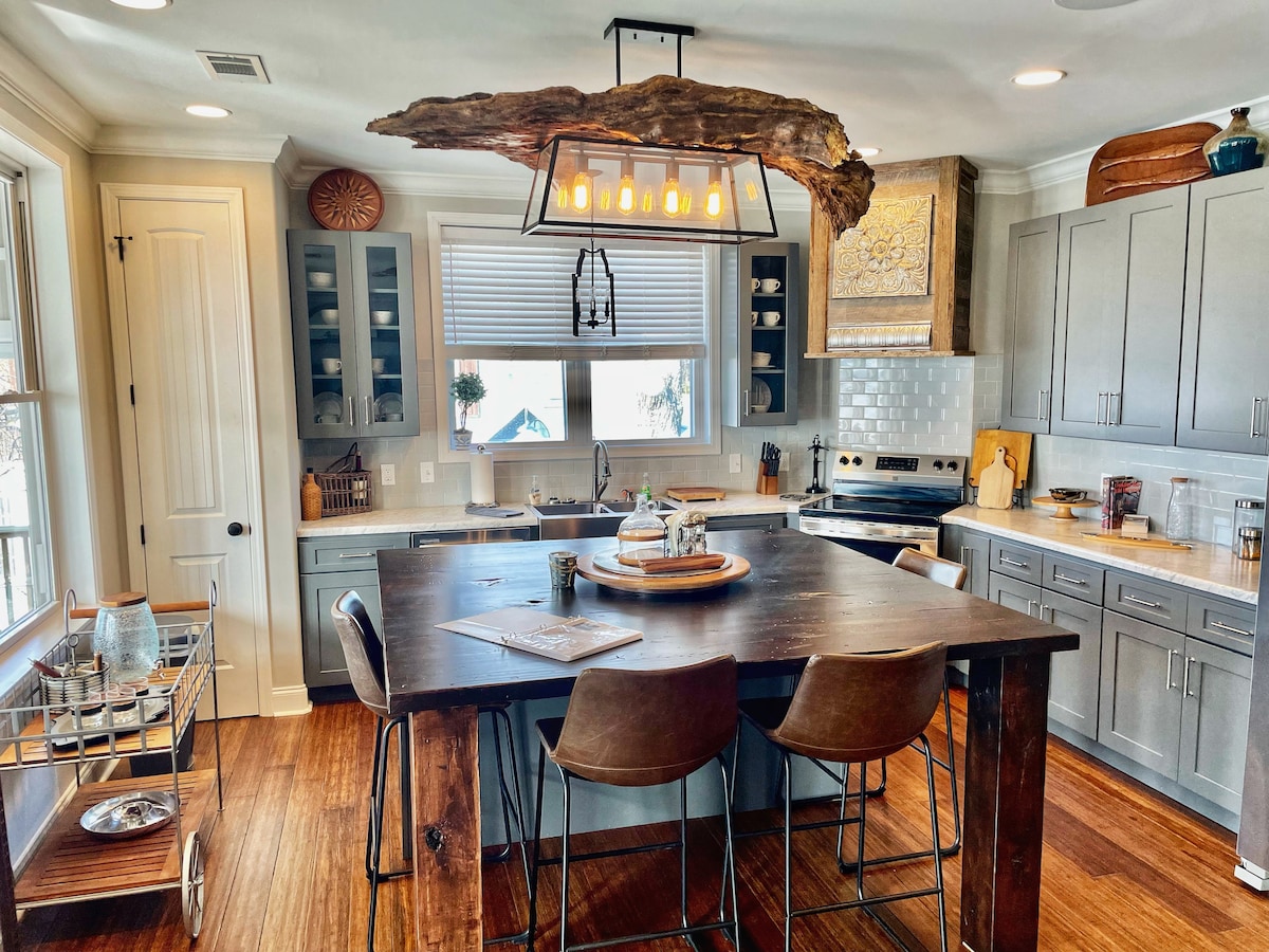 The modern kitchen showcases a large wooden dining table with four metal stools. A unique light fixture made from a natural wood branch hangs above, complementing the cabinetry. Ample counter space is available, with a variety of kitchenware visible on shelves and the countertop.