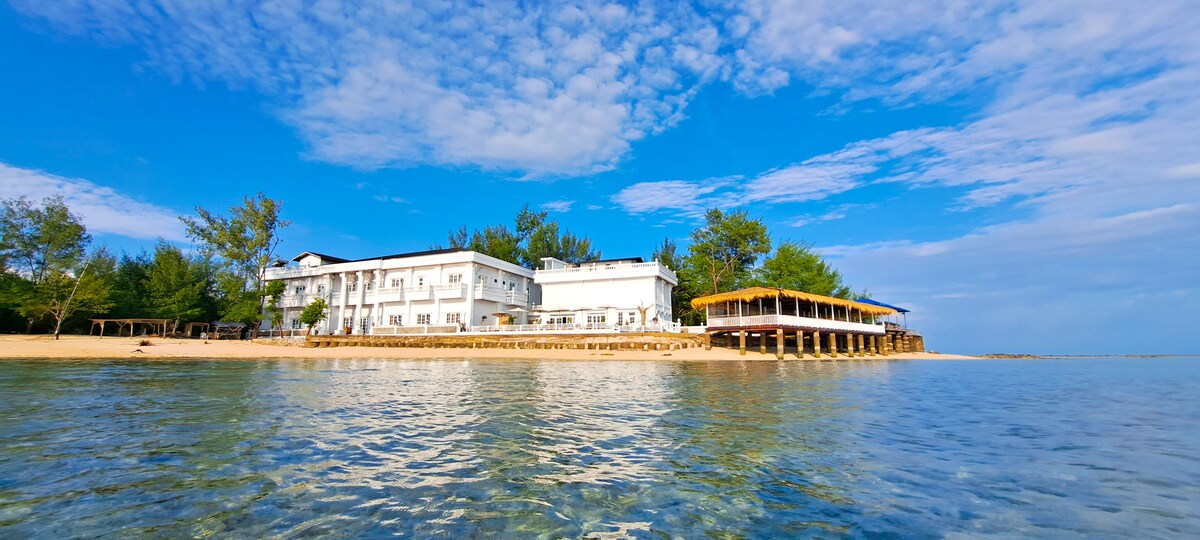 The hotel is seen along the beach, featuring a modern two-story structure with a balcony overlooking the water. The sandy shore and clear blue sky are visible with a few trees lining the beach, while gentle waves lap at the shoreline.