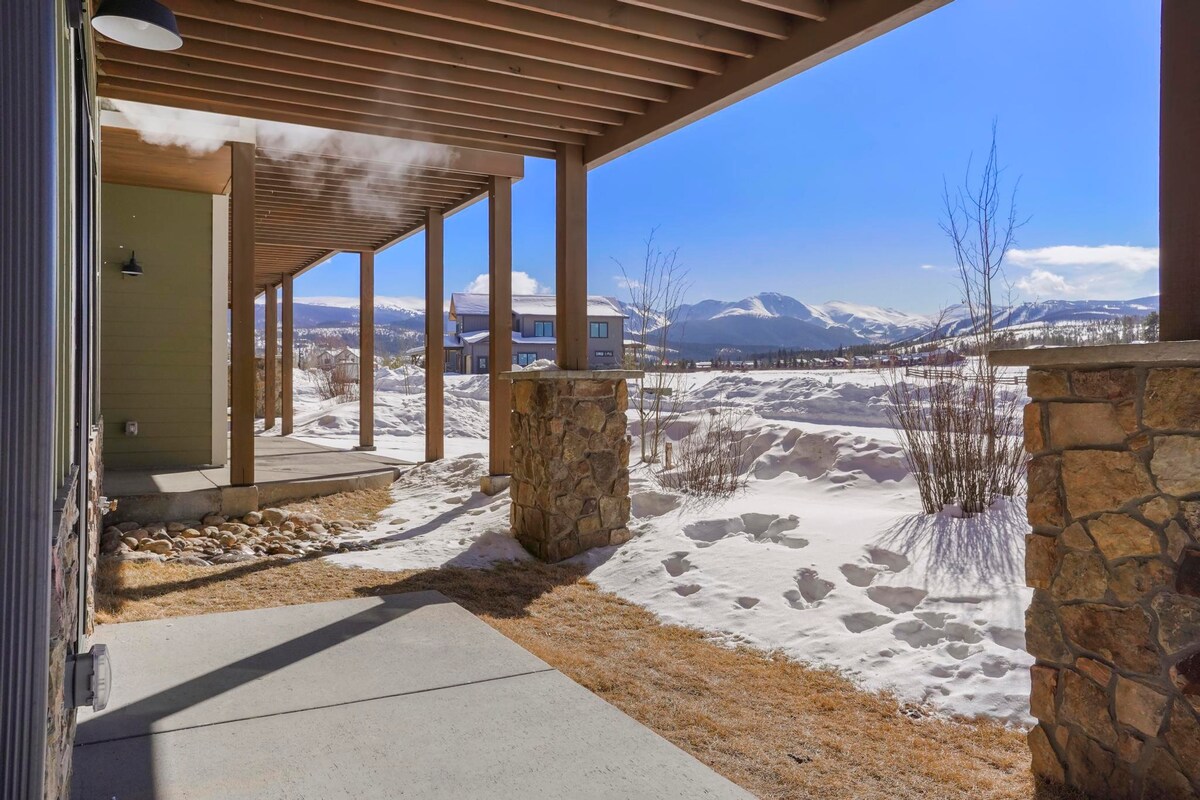 A covered porch extends alongside the building, featuring a concrete path. Snow-covered ground leads to distant mountain views under a clear blue sky. Natural stone supports the wooden beams, creating a cohesive transition between outdoor and indoor spaces.
