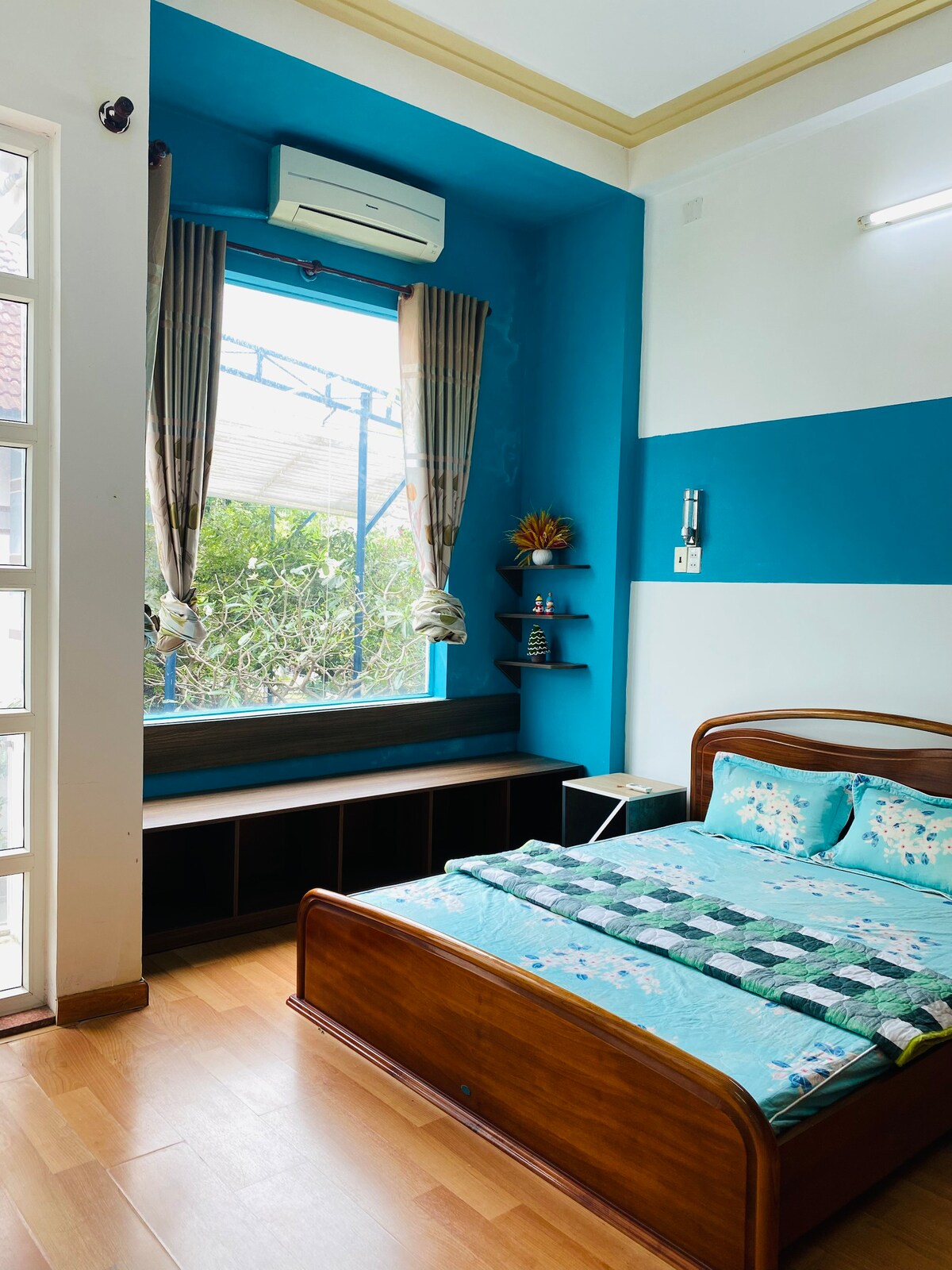 A bedroom features a wooden bed with a patterned blanket, set against a blue wall. A seating area with storage is positioned below a window, which is framed by light curtains. An air conditioning unit is installed above, providing comfort.