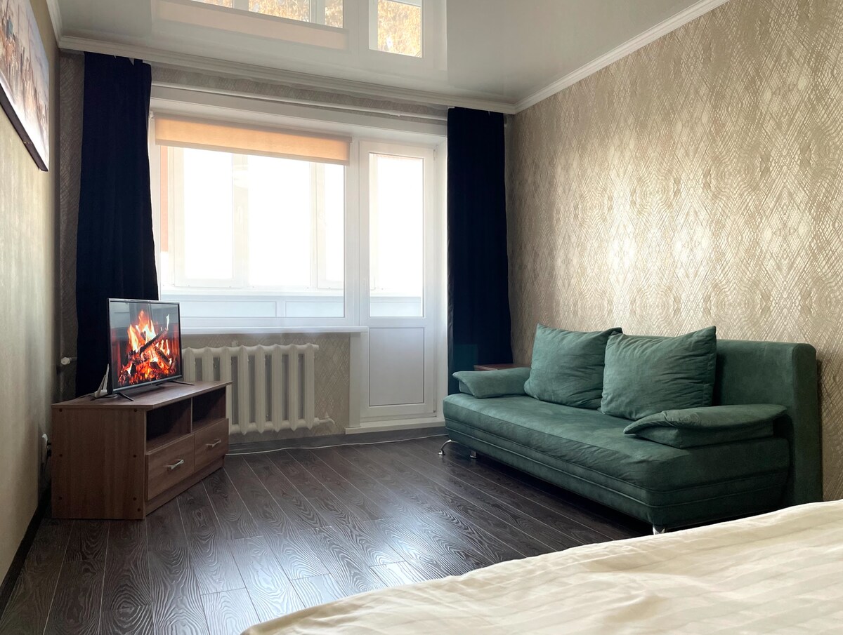 A well-lit living area is furnished with a green sofa and a wooden TV stand featuring a flat-screen television. Natural light enters through large windows covered by light shades, while dark curtains frame the window. The hardwood floor adds warmth to the space.