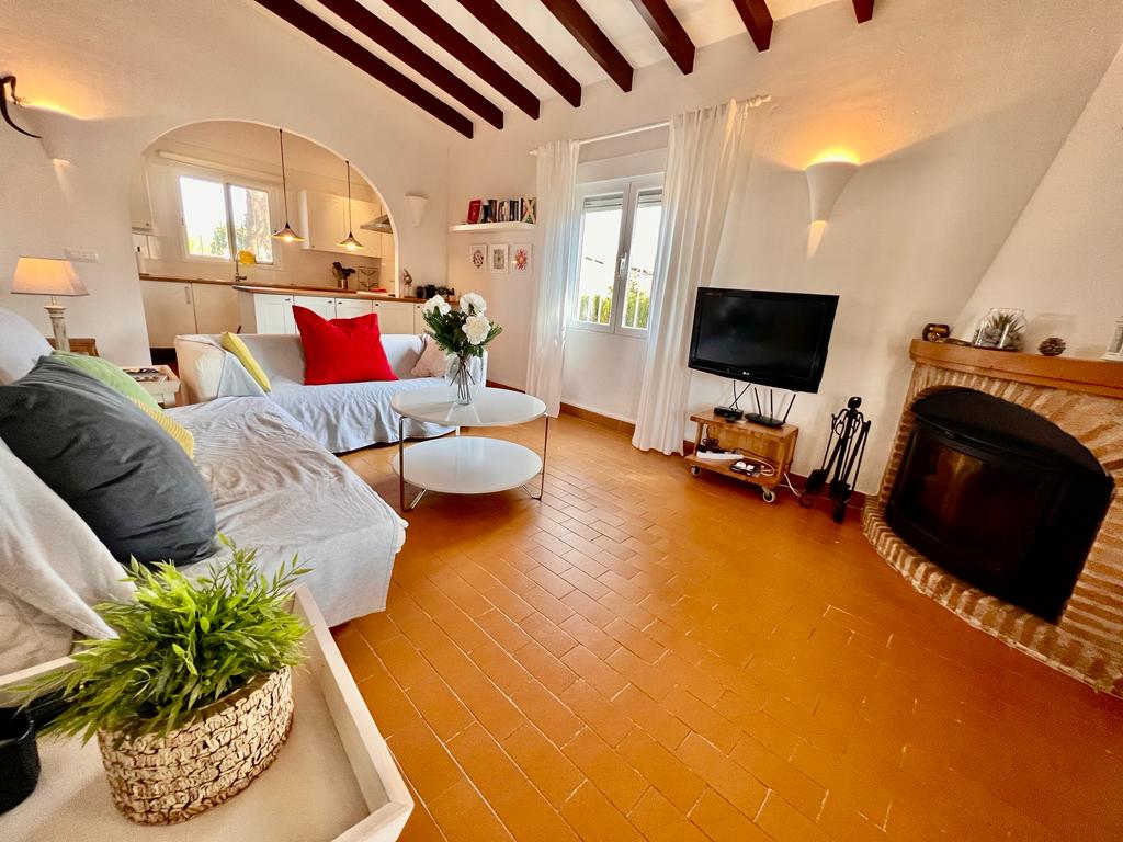 The living area features a cozy sofa with colorful cushions and a round coffee table. A television is positioned on a low stand, with natural light entering through the nearby windows. A fireplace is visible, adding warmth to the space.