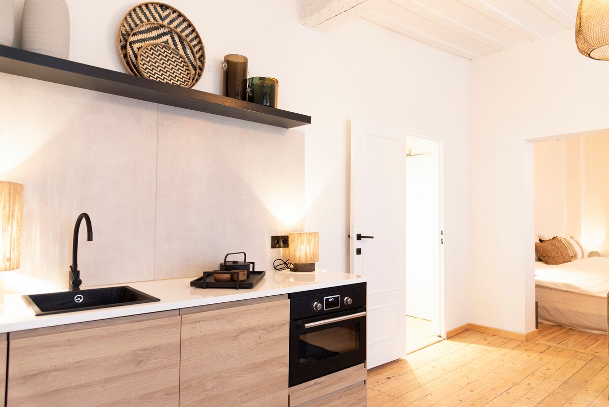 A compact kitchen features a sleek countertop with a black sink and an integrated oven below. Natural wood cabinetry adds warmth, while decorative items are placed on the shelves. Soft lighting from table lamps enhances the inviting atmosphere. A doorway leads to the bedroom area.