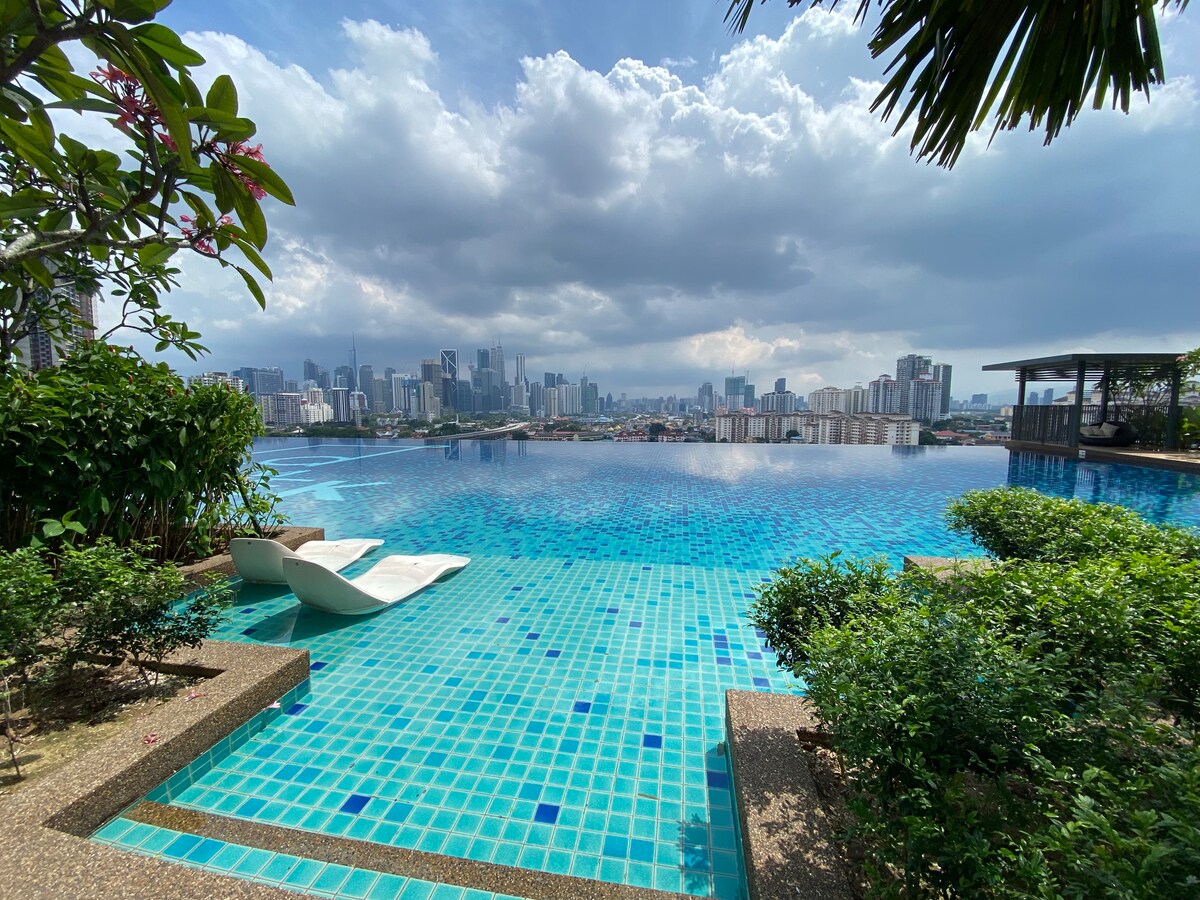 An outdoor swimming pool is visible, featuring vibrant blue tiles that reflect the sky. Loungers sit beside the pool, surrounded by lush greenery. In the background, city skyscrapers rise against a cloudy sky, creating a serene urban landscape.