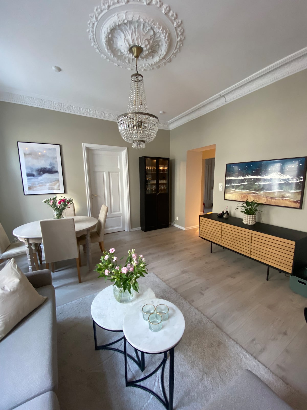 The living area features a light-colored sofa and two round coffee tables with decorative floral arrangements. A dining table with upholstered chairs is visible, along with a sideboard and an artwork hanging on the wall. Natural light enters through the doorway, enhancing the calm atmosphere.