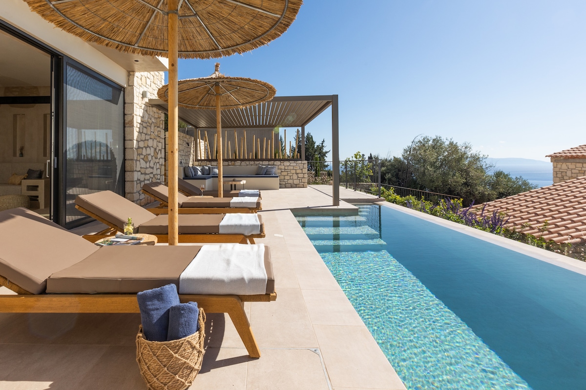 A sunlit terrace features wooden loungers arranged beside a tranquil private pool. The area is shaded by thatched umbrellas, and a spacious outdoor seating structure is visible in the background, providing a clear view of the surrounding landscape and distant sea.