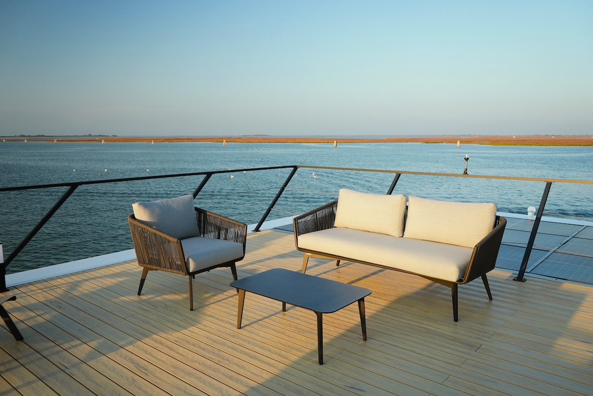 A comfortable outdoor seating area features a sofa and two armchairs arranged around a small table, set on a wooden terrace. The scene is accentuated by scenic water views, with a clear sky and a tranquil atmosphere enhancing the experience of relaxation.