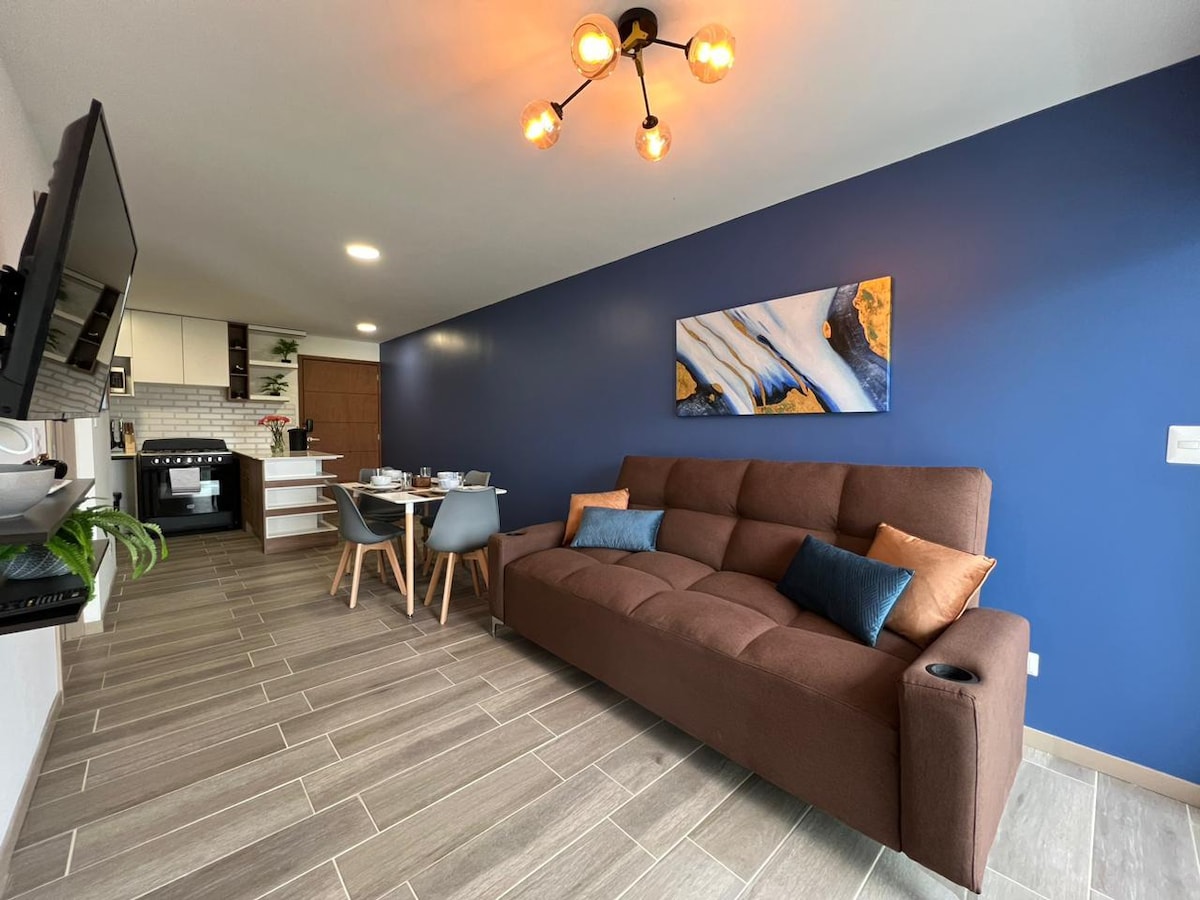 A modern living area features a spacious brown sofa adorned with decorative pillows. A dining table with grey chairs is positioned near the open kitchen, which showcases sleek cabinetry and appliances. Natural light complements the blue accent wall, enhancing the inviting ambiance.