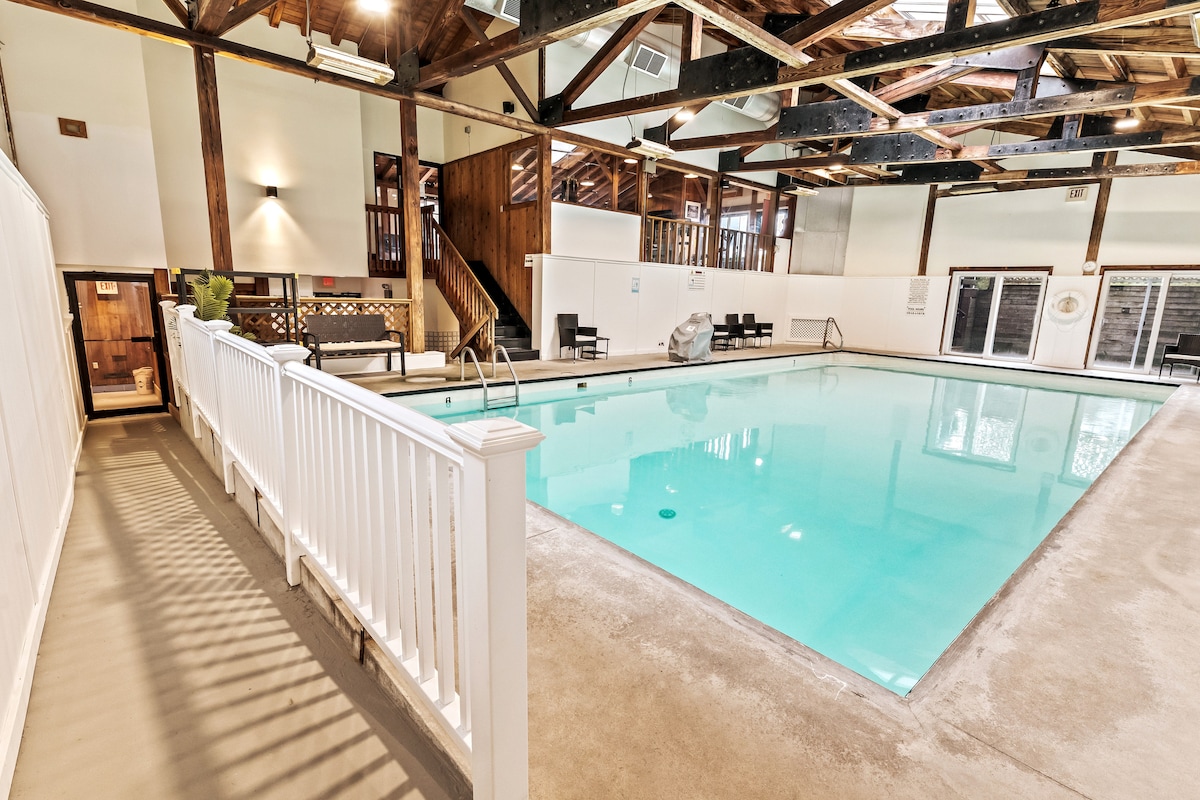 An indoor heated pool is showcased, featuring clear blue water under a spacious wood-beamed ceiling. Lounge chairs line the sides, providing a place to relax. Natural light filters in through large windows, creating a bright and inviting atmosphere.