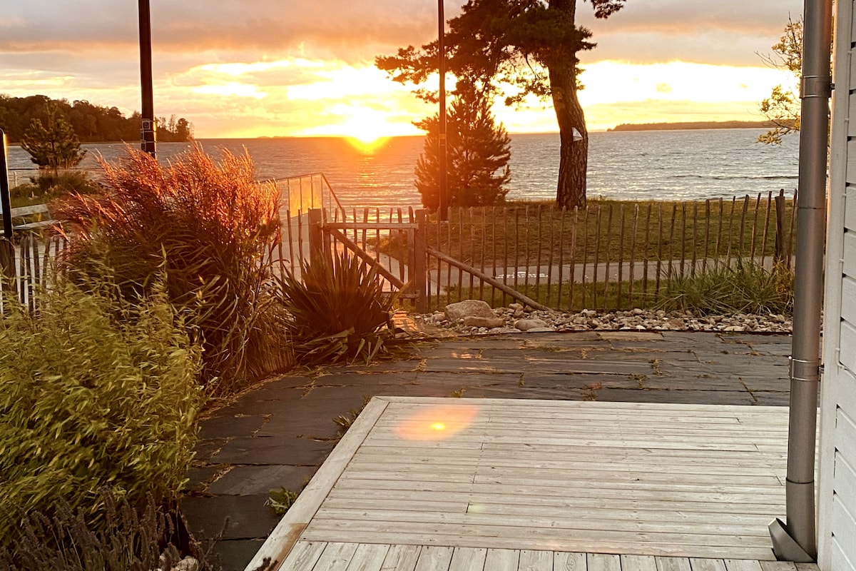 A view of a sunset over the water is captured from a wooden patio area. Soft grasses and shrubs frame the scene, while a pathway and a wooden fence lead towards the shoreline. The sky is painted in warm hues, creating a serene backdrop.